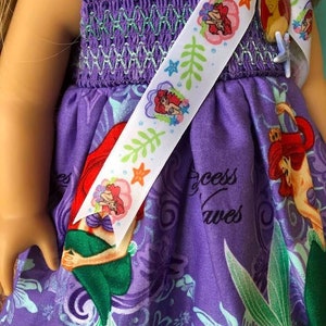 Op de afbeelding: Een paarse poppenjurk met een zeemeerminprint en een witte lint sjerp. De jurk heeft een gesmokt lijfje met groene stiksels en een rok met een herhalend patroon van zeemeerminnen, zeewier en de woorden "Princess Waves". De sjerp is versierd met zeemeermin illustraties.