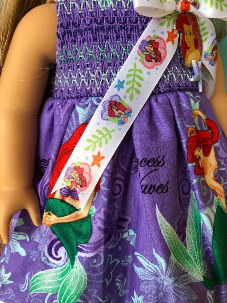 Op de afbeelding: Een paarse poppenjurk met een zeemeerminprint en een witte lint sjerp. De jurk heeft een gesmokt lijfje met groene stiksels en een rok met een herhalend patroon van zeemeerminnen, zeewier en de woorden "Princess Waves". De sjerp is versierd met zeemeermin illustraties.