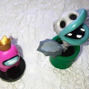 May include: Two figurines on a white lace surface. One is a pink character with a gold crown, black base, and a blue visor. The other is a plant-like figure with a white skull-like head, teal mouth, and a green pot.