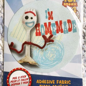 May include: Round, light blue adhesive fabric featuring the Toy Story character Forky with the text "I'M HOMEMADE" in red and blue. The packaging includes the Disney Pixar logo and the words "PEEL & STICK".