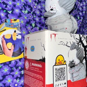 May include: A collectible rubber duck figure styled as Pennywise from IT Chapter Two, featuring grey hair, a ruffled collar, and red blood splatters. The figure sits atop its white and red cardboard display box, which includes a QR code and text.