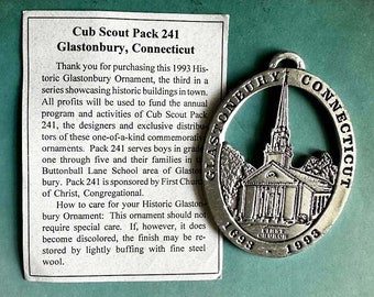 May include: Silver-toned ornament featuring a church building with the text "GLASTONBURY CONNECTICUT 1698 CHURCH 1993".