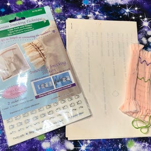 May include: A craft image featuring a Clover Nouveau Smocking Technique kit with stickers, instructions, and a pink fabric sample. The kit includes a guide in multiple languages. The fabric sample has green and purple stitching.