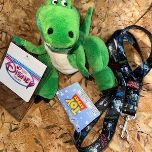 May include: A green plush Rex dinosaur toy with a white belly, black eyes, and a tag from The Disney Store. A Toy Story lanyard with Buzz and Woody characters is also present.