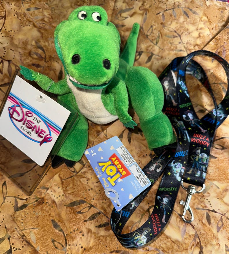 May include: A green plush Rex dinosaur toy with a white belly, black eyes, and a tag from The Disney Store. A Toy Story lanyard with Buzz and Woody characters is also present.