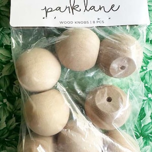 May include: A package of eight unfinished wood knobs. The package has a white label with the words "park lane" in cursive and "WOOD KNOBS | 8 PCS" in a smaller font. The knobs are light brown and have a rounded shape.