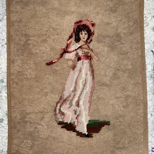 May include: Needlepoint tapestry featuring a woman in a pink and white dress and a matching bonnet. The artwork depicts a figure with dark hair and a ribbon, set against a tan background. The tapestry is a vintage piece, showcasing a classic design.