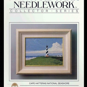 May include: A framed needlework art piece titled National Park Needlework Collector Series featuring the Cape Hatteras National Seashore. The image depicts a black and white striped lighthouse on a green grassy dune under a blue sky with clouds.