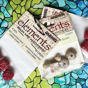 May include: Two packages of beads and accessories. The first package contains 4 carved Asian beads in a red color. The second package contains 4 beads in a light brown color. The packaging is labeled "elements beads and accessories" and "the beadery craft products".