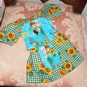 May include: A green and white gingham dress with a blue and white applique featuring the characters from the movie Frozen. The applique has a blue background with white snowflakes and stars. The dress has a green and white gingham skirt with sunflowers printed on it.