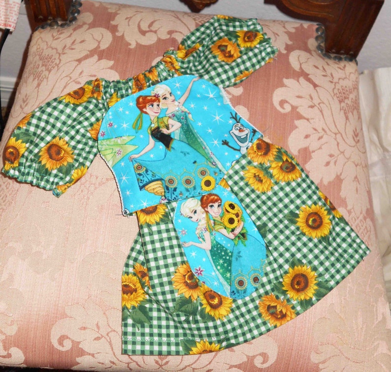 May include: A green and white gingham dress with a blue and white applique featuring the characters from the movie Frozen. The applique has a blue background with white snowflakes and stars. The dress has a green and white gingham skirt with sunflowers printed on it.