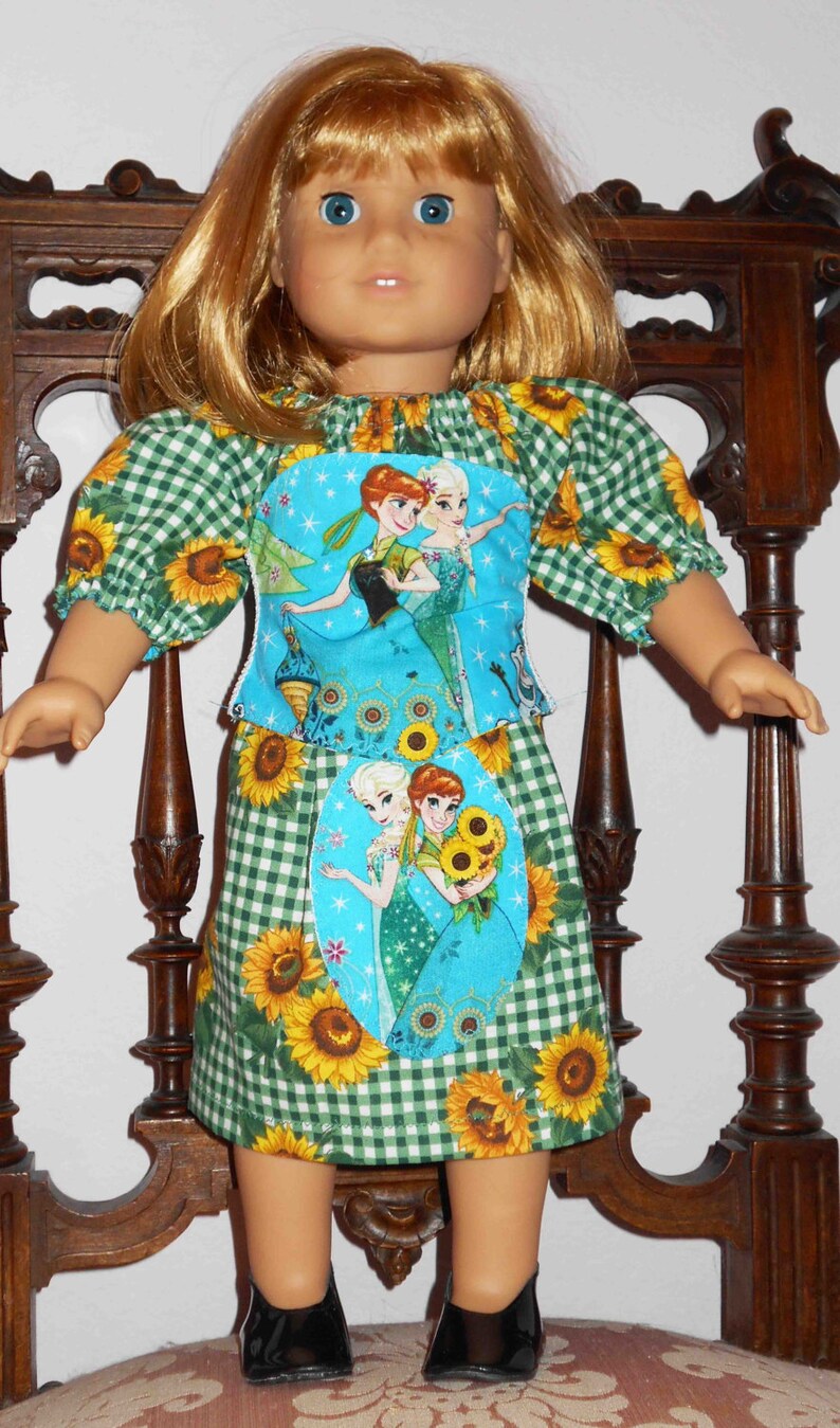 May include: A doll wearing a green and white gingham dress with a blue and white fabric panel featuring a scene from the movie Frozen. The dress has sunflowers printed on it. The doll is wearing black shoes.