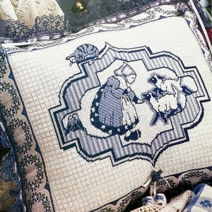 May include: A rectangular decorative pillow featuring a cross-stitch design of a person in a bonnet tending to two sheep. The embroidery is in shades of blue on a cream fabric, framed by a dark blue lace border with small floral patterns.