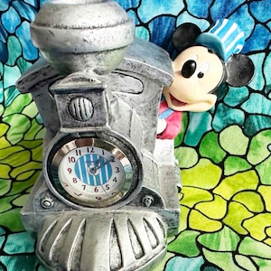 May include: A silver-colored train engine clock with a Mickey Mouse figurine peeking out from the side. The clock face has blue and white stripes and the words "Mickey Mouse" printed on it.