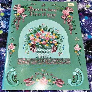 May include: A Bavarian Heritage book cover with a floral basket design. The cover is green with a central light blue arch containing a basket of pink, yellow, and blue flowers. The title is in ornate lettering, and the artist's name is at the bottom.