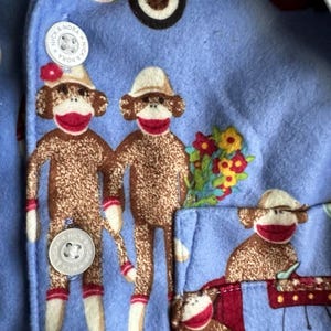 May include: Blue pajama shirt with a pattern of monkeys wearing red socks and hats. The shirt has a pocket on the left side and two buttons with the text "Nick & Nora" on them.