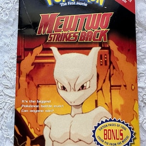 May include: A book cover featuring a white Pokémon character with red eyes, against a fiery orange background. The title is "Pokémon: The First Movie Mewtwo Strikes Back".