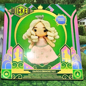 May include: A Wicked Glinda crochet kit box featuring a crocheted doll with blonde hair and a pink dress. The packaging includes the text Wicked, Glinda Crochet Kit, and lists contents including 5 colors of yarn, 148.73 yards total, and 0.42 ounces of stuffing.