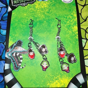 May include: A pair of silver-toned dangle earrings featuring cartoon character charms. The charms depict characters from the movie "Beetlejuice" on a green background. The packaging includes the text "Beetlejuice" and "Exclusive Claire's".