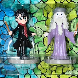 May include: Four figurines from the Harry Potter series. The figures include Ron Weasley, Harry Potter, Dumbledore, and Hermione Granger. Each figure is holding a wand and is on a silver base. The background is a colorful mosaic.