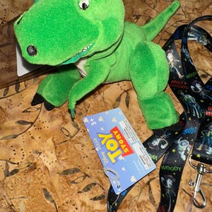 May include: A green plush Rex dinosaur toy from the movie Toy Story, with a blue and white tag. A black lanyard with Woody and Buzz Lightyear characters is also present.