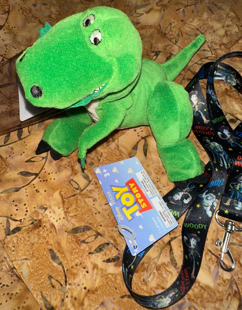 May include: A green plush Rex dinosaur toy from the movie Toy Story, with a blue and white tag. A black lanyard with Woody and Buzz Lightyear characters is also present.