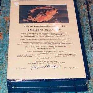 May include: A VHS tape titled Mollusks in Action, featuring a beige cover with a photograph of a seashell. The text describes a 30-minute documentary about living seashells filmed on Sanibel Island, Florida, produced by Joyce Matthys in 2003.