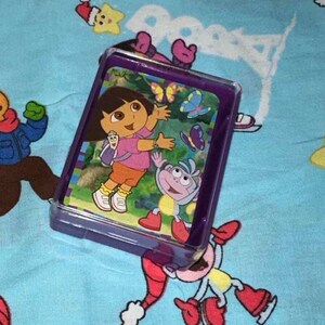 May include: A purple plastic case with a picture of Dora the Explorer and Boots the monkey on the front. Dora is wearing a purple shirt and a backpack. Boots is wearing a blue hat and a red shirt. There are butterflies in the background.