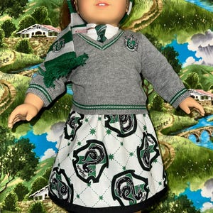 May include: A doll wearing a gray sweater with green trim, a matching scarf, and a skirt featuring a repeating pattern of a green serpent on a white background. The doll's outfit is inspired by a popular fantasy series.