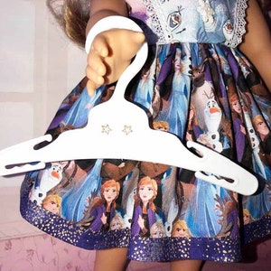 K&ouml;nnte beinhalten: Puppe in einem Frozen-Themenkleid mit einem wei&szlig;en Kleiderb&uuml;gel. Das Kleid zeigt Charaktere wie Anna, Elsa und Olaf, mit einem blau-violetten Farbschema. Das Kleid hat eine wei&szlig;e Spitzenborte und den Text "Ice Breaker!"