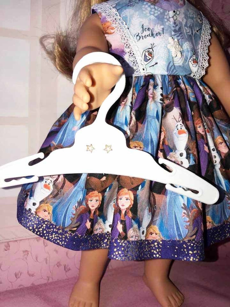 K&ouml;nnte beinhalten: Puppe in einem Frozen-Themenkleid mit einem wei&szlig;en Kleiderb&uuml;gel. Das Kleid zeigt Charaktere wie Anna, Elsa und Olaf, mit einem blau-violetten Farbschema. Das Kleid hat eine wei&szlig;e Spitzenborte und den Text "Ice Breaker!"