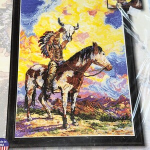 May include: A framed counted cross-stitch picture kit featuring a Native American on horseback holding a skull. The artwork depicts a colorful sunset and mountains. The kit measures 12 x 18 inches and includes floss and instructions.