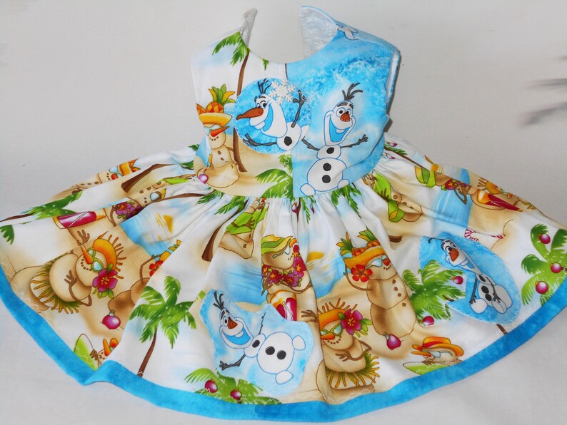 Pode incluir: Um vestido branco sem mangas com uma bainha azul. O vestido apresenta uma estampa tropical com palmeiras, bonecos de neve e os personagens Olaf e Sven do filme Frozen.