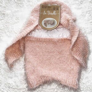 May include: A light pink newborn photography outfit with a hood and attached bunny ears. The outfit is made of a soft, fuzzy material. A tag reads "So Doralle" with a photo of a baby.