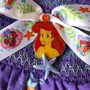Op de afbeelding: Close-up van een paarse jurk met een wit lint met Ariel van De Kleine Zeemeermin. Het lint is versierd met illustraties met zeethema. Een Ariel-speld en een zeester-bedel zijn bevestigd.