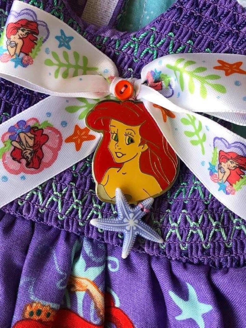 Op de afbeelding: Close-up van een paarse jurk met een wit lint met Ariel van De Kleine Zeemeermin. Het lint is versierd met illustraties met zeethema. Een Ariel-speld en een zeester-bedel zijn bevestigd.