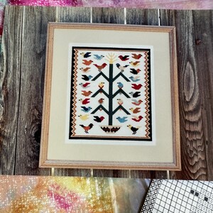 May include: Framed cross-stitch artwork featuring a tree with colorful birds. The birds are in various colors, including red, blue, yellow, and black. The artwork is framed in a light wood frame with a cream-colored mat.