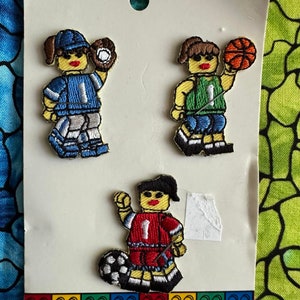 May include: Three embroidered iron-on patches featuring cartoon figures playing sports. The patches are on a white card with a Lego logo at the bottom. The figures are a baseball player, a basketball player, and a soccer player.