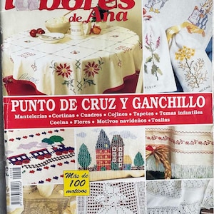 May include: A magazine cover featuring a colorful cross-stitch design of a house with a red roof and green trees. The magazine title is "Las Labores de Ana" and the text "Punto de Cruz y Ganchillo" is below the image. The magazine cover also features other cross-stitch designs, including a train, a table cloth, and a towel.