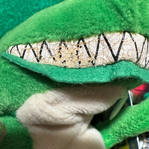 May include: Close-up of a green plush toy with a detailed, textured mouth and teeth. The plush has a white underbelly and a green, fuzzy exterior. The teeth are off-white with black zigzag lines.