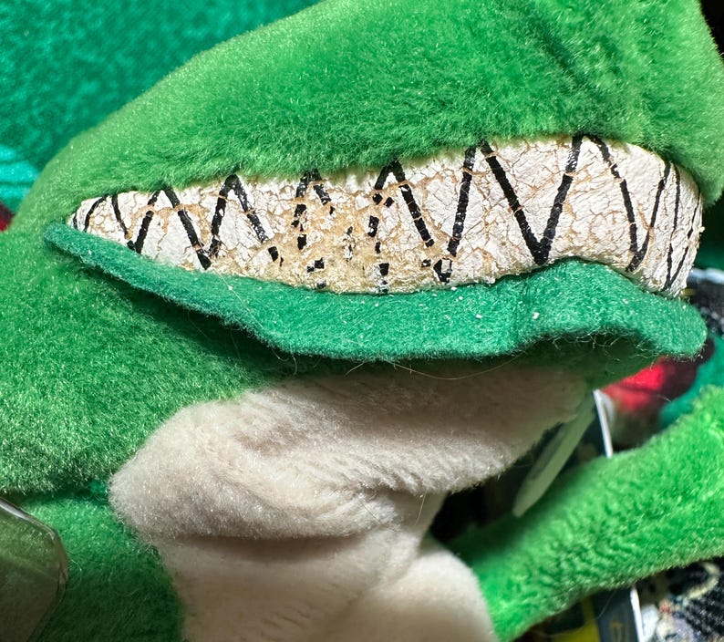 May include: Close-up of a green plush toy with a detailed, textured mouth and teeth. The plush has a white underbelly and a green, fuzzy exterior. The teeth are off-white with black zigzag lines.