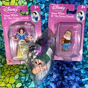 May include: Two Disney Princess Snow White and the Seven Dwarfs figurines in their original packaging. One figurine is Snow White in a blue and yellow dress, and the other is a dwarf in an orange and blue outfit. The packaging is pink.