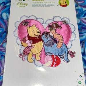 May include: A Disney baby appliqué featuring Winnie the Pooh, Piglet, Eeyore, and Tigger. The characters are embroidered on a pink heart-shaped patch with a scalloped edge. The text "Simplicity" and "Appliqué" are visible.