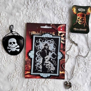 May include: A set of Pirates of the Caribbean themed accessories including a black eye patch with a white skull and crossbones, a silver chain necklace with a heart pendant, and a black and white patch featuring a character from the movie.