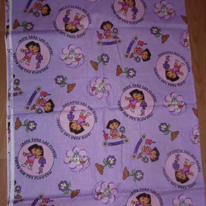 May include: Lavender fabric featuring a repeating pattern of Dora the Explorer, flowers, and the text "Agua Para Las Flores! Water for the Flowers!". The design includes Dora with a watering can and other floral elements.