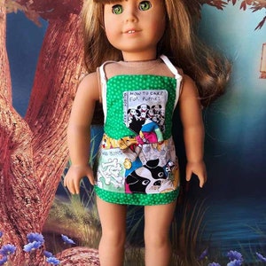 May include: A doll wearing a green and brown apron with a book design. The book cover says "How to Care for Puppies".