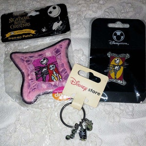 May include: A set of Nightmare Before Christmas themed merchandise featuring a pink pillow-shaped iron-on patch, a black and red pin with Jack Skellington, and a silver keychain with Jack Skellington charms. The packaging includes the Disney store logo.