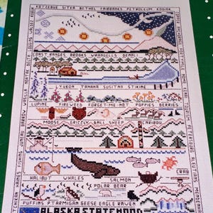 May include: Cross-stitch sampler depicting Alaskan landmarks, wildlife, and flora. The design includes text such as "Alaska Statehood 1959-1984" and various place names. The color palette features blues, greens, and browns, with detailed stitching creating a textured effect.