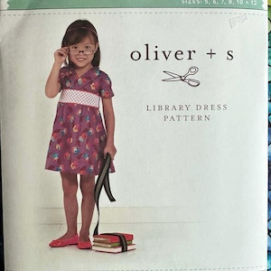 May include: A sewing pattern for a girl's dress, called "Library Dress" by Oliver + S. The pattern is for sizes 5, 6, 7, 8, 10, and 12. The image shows a young girl wearing a purple and white dress with a checkered waistband, holding a stack of books.