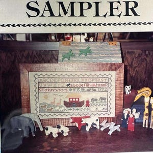 May include: A framed cross-stitch sampler with the title "Noah's Ark Sampler". The sampler features the alphabet, numbers, and a depiction of Noah's Ark with animals. The frame is made of wood and has a light brown finish. The sampler is surrounded by a variety of wooden animal figurines, including a giraffe, elephant, cow, and rabbit.